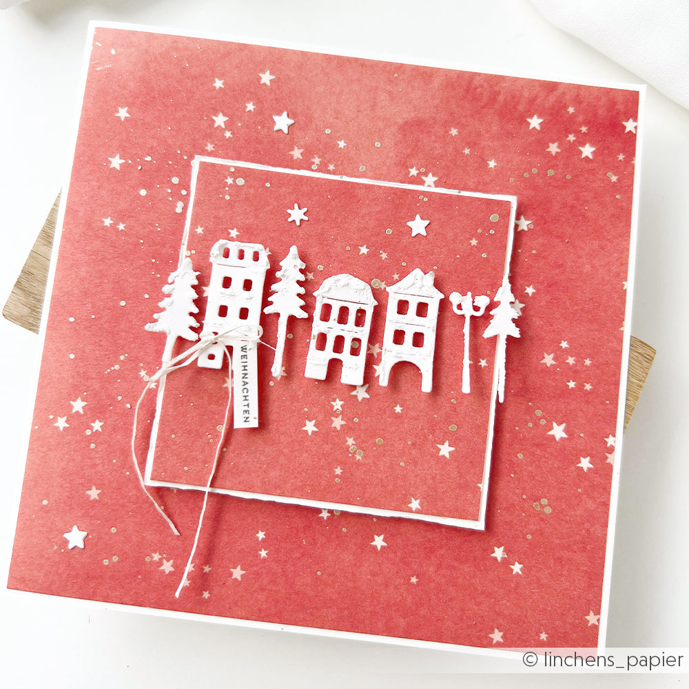 Handgefertigte, leuchtend rote Weihnachtskarte mit weißen, ausgestanzten Häusern, Bäumen und kleinen Sternen.