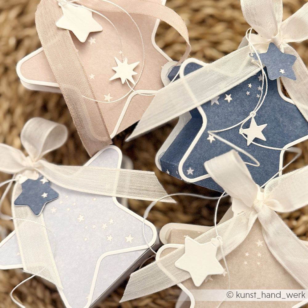 Vier sternförmige Geschenkboxen aus Designpapier, verziert mit Bändern und kleinen Stern-Anhängern.