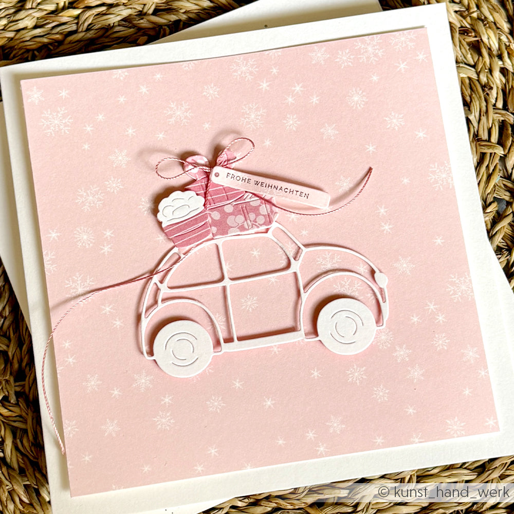 Handgefertigte rosa Weihnachtskarte mit einem ausgestanzten Auto, das ein Geschenk transportiert, und einem Anhänger mit der Aufschrift „FROHE WEIHNACHTEN“ auf Papier mit Schneeflockenmuster.