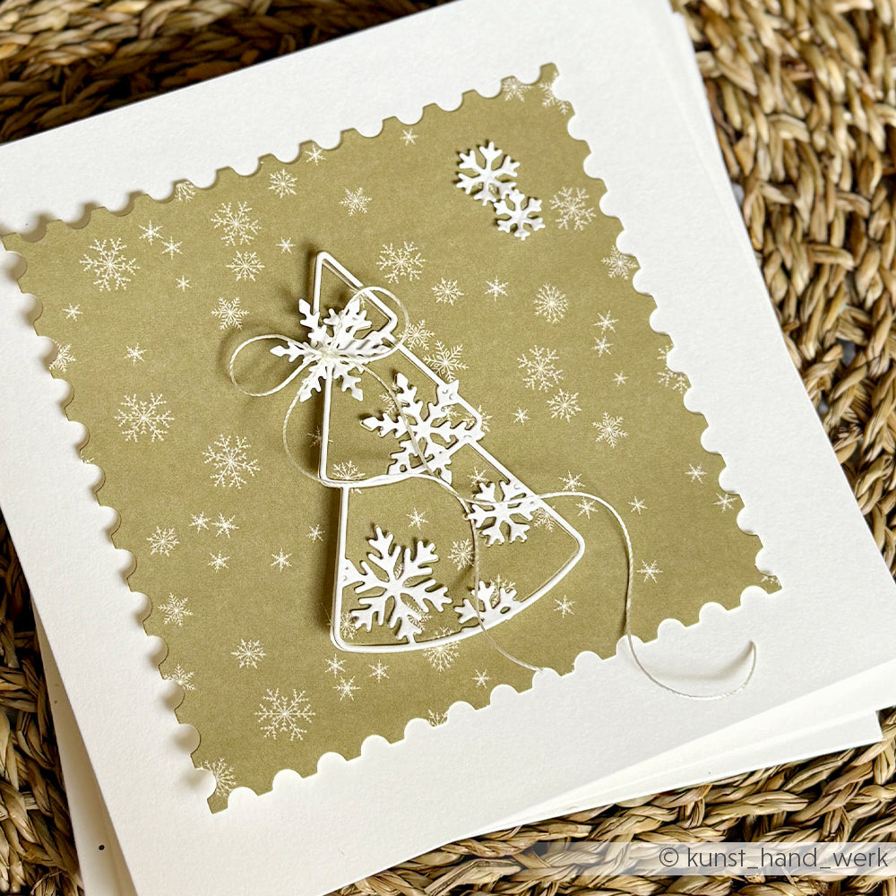 Eine handgefertigte Weihnachtskarte mit einem stilisierten Drahtbaum, der mit weißen Schneeflocken-Anhängern geschmückt ist, auf gemustertem Designpapier in sanften Grüntönen.