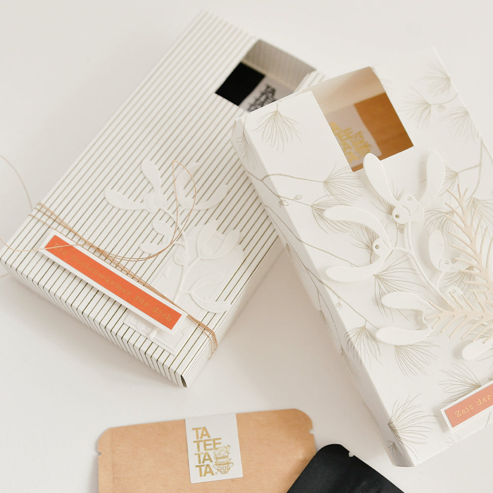 Zwei Geschenkboxen, verpackt in Designpapier mit dem Muster „Zarte Streifen Schattengrün“, verziert mit Mistelzweigen und einem kleinen Päckchen mit der Aufschrift „Ta Tee Ta Ta“.