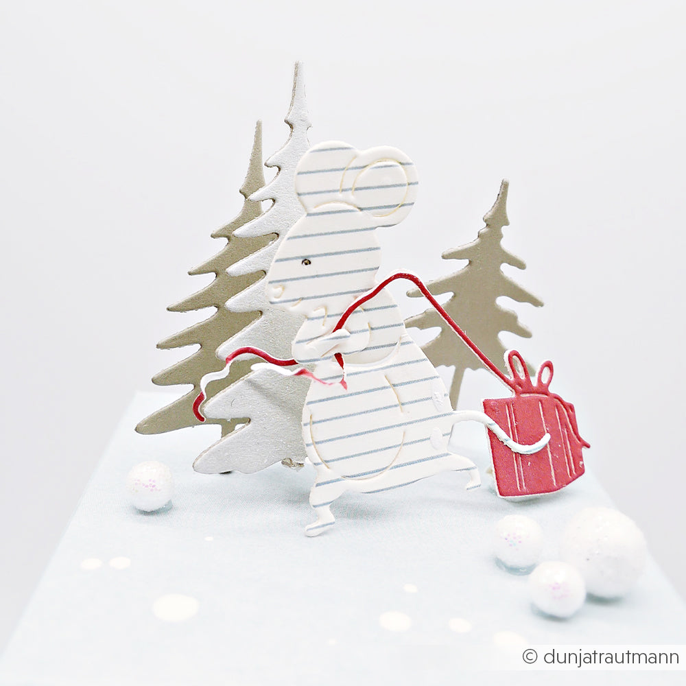 Eine aus Papier ausgeschnittene Maus zieht ein rotes Geschenk durch einen verschneiten Wald – gestaltet als Papierbastelprojekt.