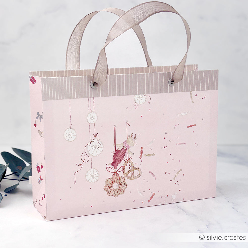 Rosa Geschenktüte aus Designpapier mit einem Rentier auf einer Schaukel und hängenden Weihnachtskugeln.