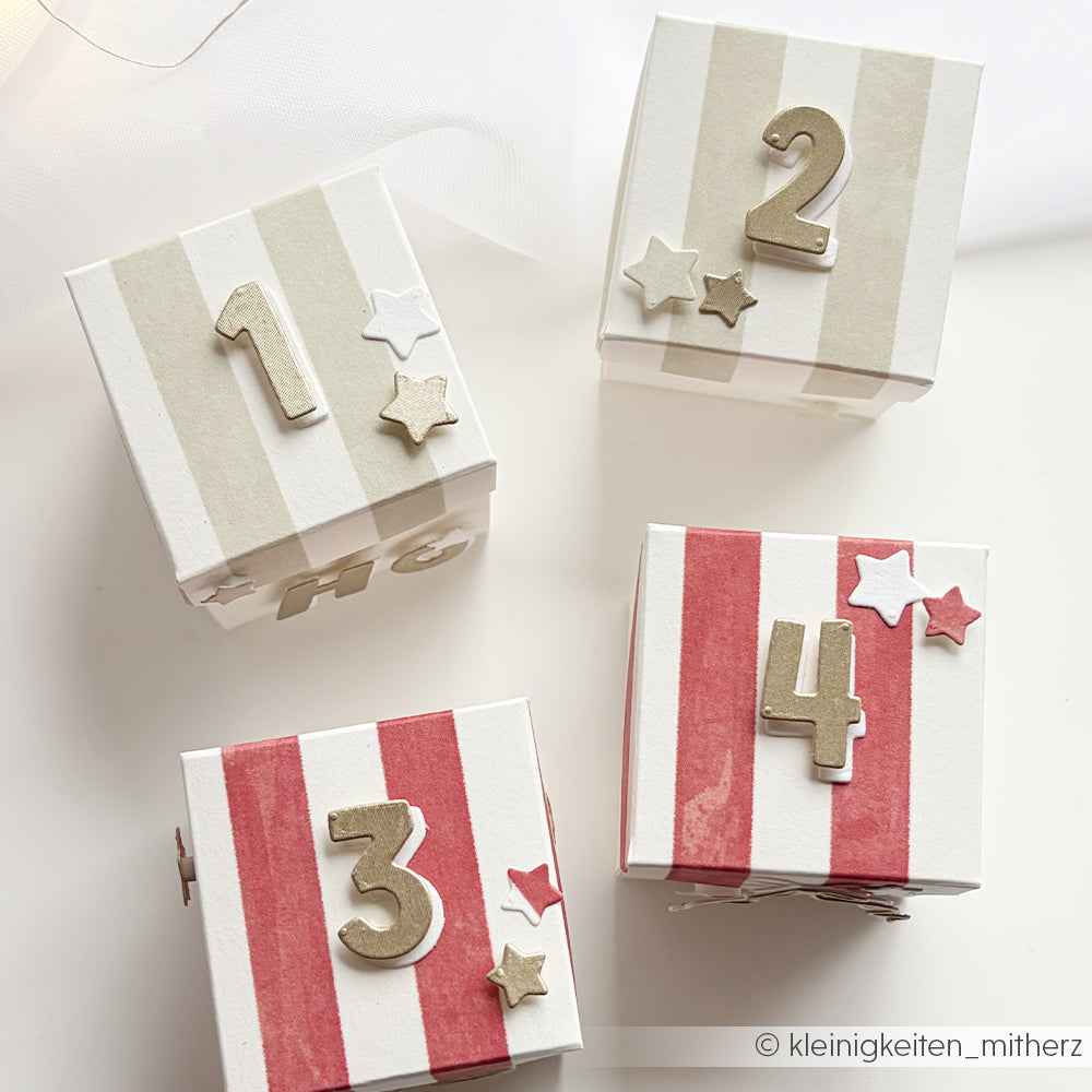 Vier kleine, mit Wasserfarben gestreifte Geschenkboxen, nummeriert von eins bis vier und mit Sternen verziert.
