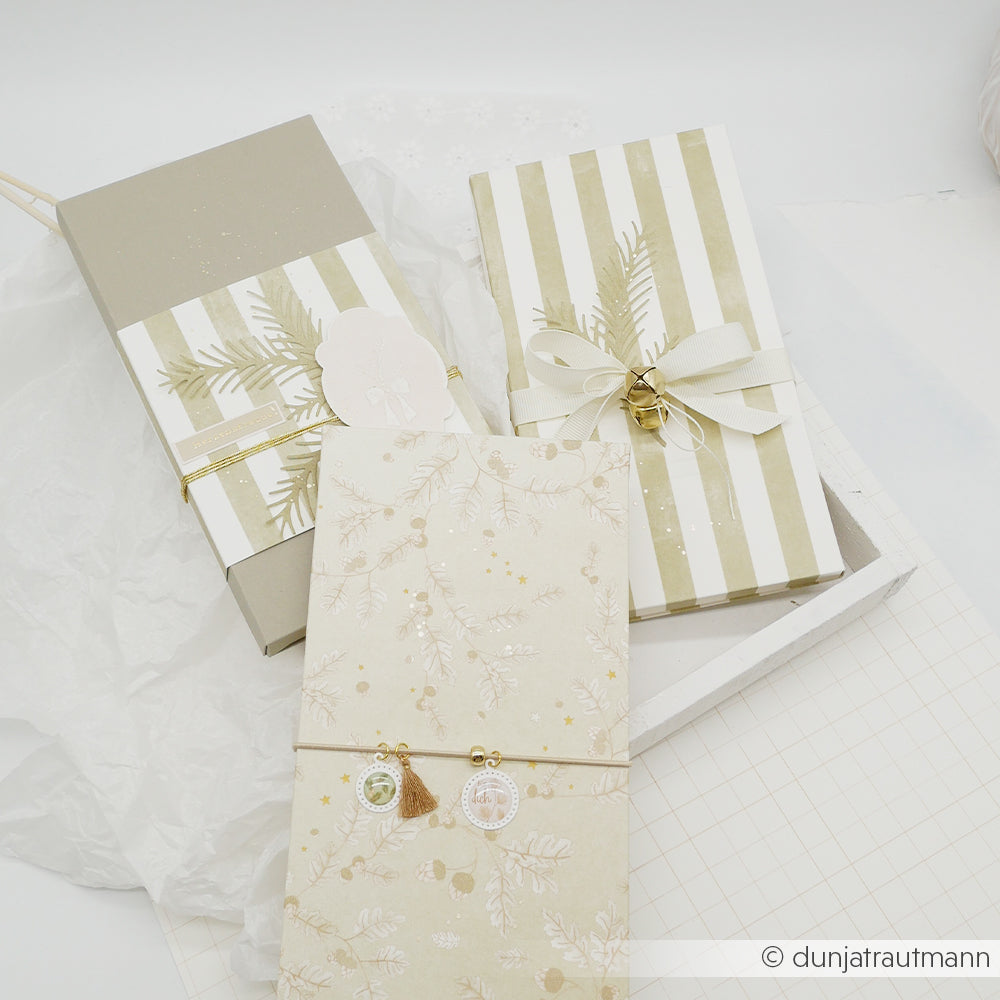 Drei Geschenkboxen, verpackt in moosgrünem, mit Wasserfarbenstreifen bedrucktem Papier, verziert mit Bändern und weihnachtlichen Anhängern.
