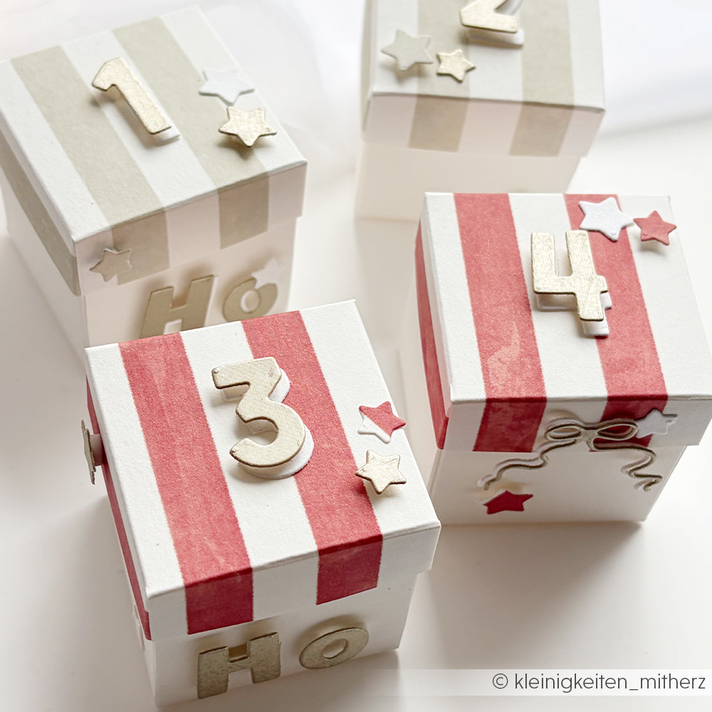 Vier quadratische Geschenkboxen in Premiumrot und mit grauen Aquarellstreifen, mit Sternausschnitten und Zahlen.