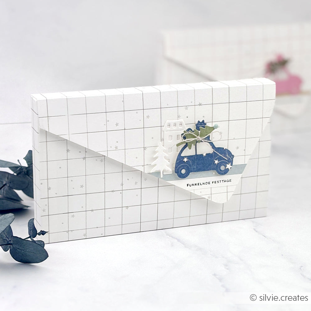 Weißer Umschlag aus Designpapier mit Gittermuster, auf dem ein blaues Auto, ein Haus und der Schriftzug FUNKELNDE FESTTAGE ausgestanzt sind.
