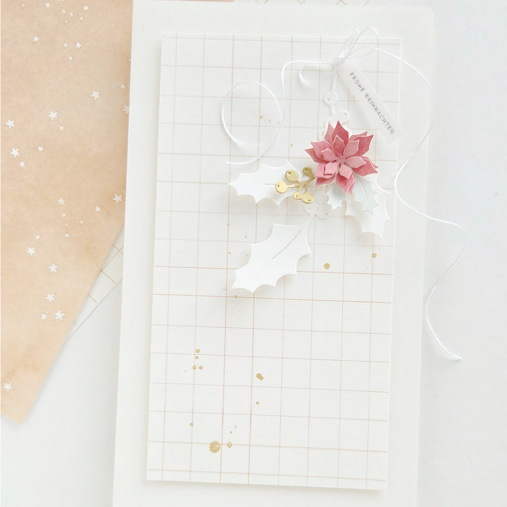 Eine Weihnachtskarte mit Karomuster, verziert mit einer Papier-Weihnachtssternblüte, Stechpalmenblättern und einem „Frohe Weihnachten“-Anhänger, die handgefertigte Papierprojekte präsentiert.