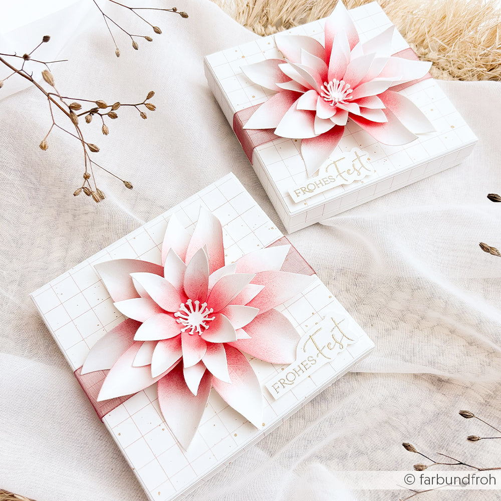 Zwei quadratische Geschenkboxen mit rosa kariertem Designpapier, Papierblumen und der Aufschrift Frohes Fest.