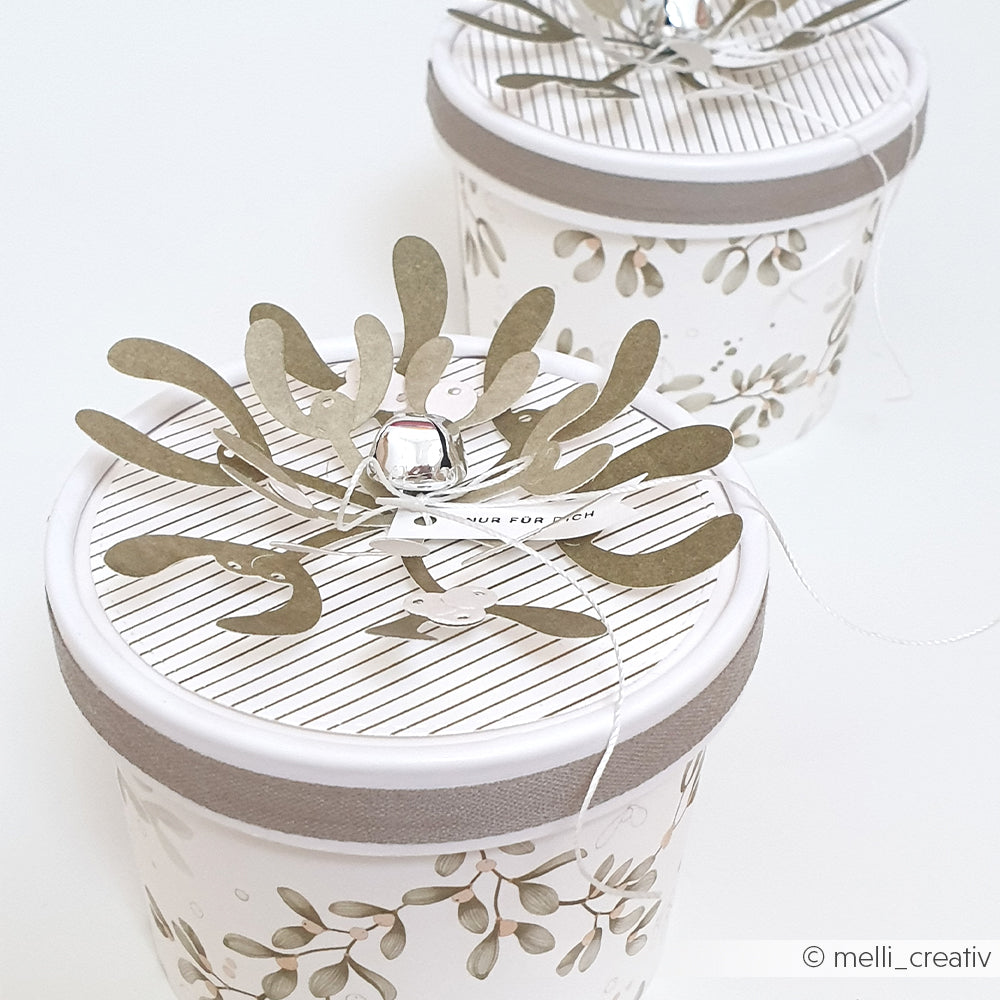 Zwei runde Mimi Moosgrün-Geschenkboxen mit Bastelprojekten aus Mistelzweig-Papier und Papierzweigen.