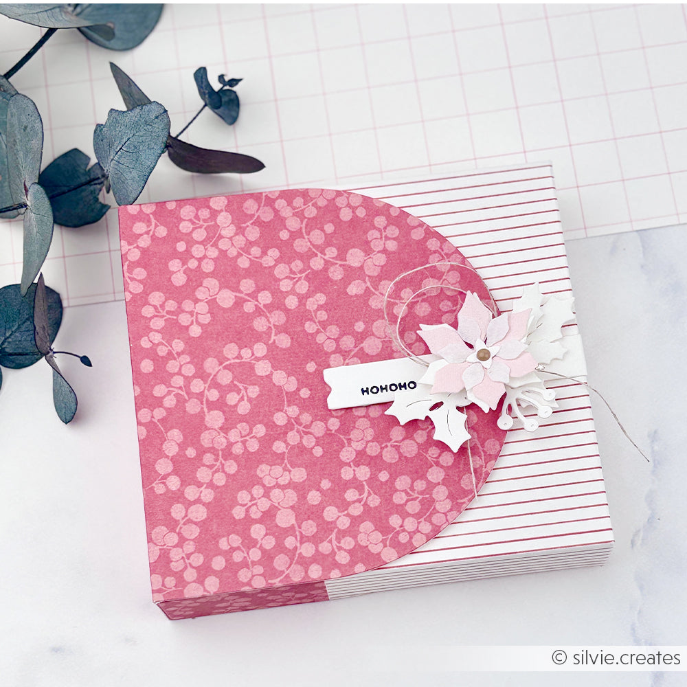 Quadratische Geschenkbox, verpackt in hochwertigem rotem Designpapier mit einem Weihnachtsstern und einem HOHOHO-Anhänger.