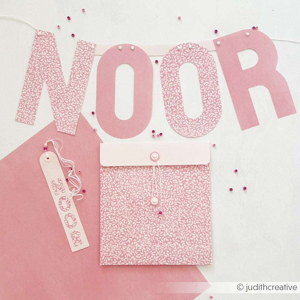 Rosa Banner mit der Aufschrift NOOR über einem passenden Lesezeichen und einem Umschlag für Papierprojekte.