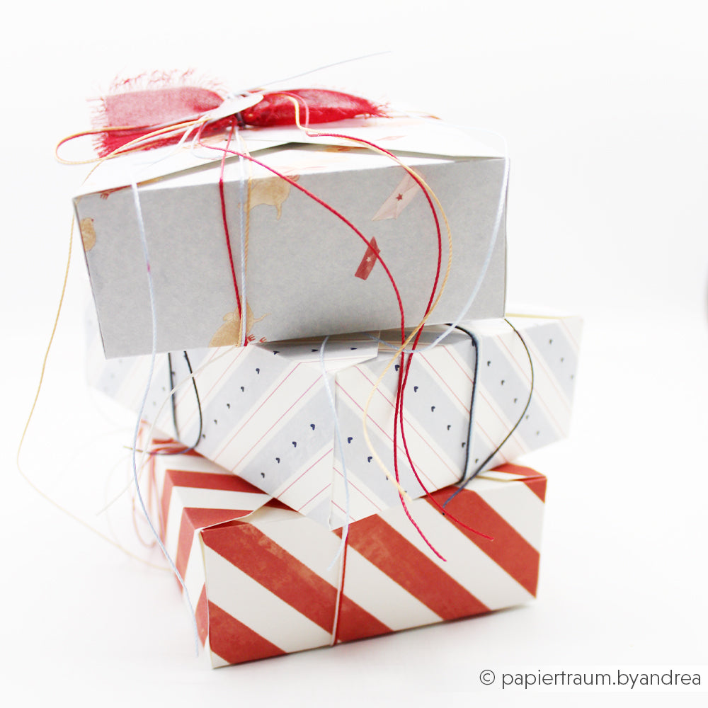 Drei übereinander gestapelte Geschenkboxen, in Designpapier eingewickelt und mit einer dünnen, bunten Schnur zusammengebunden.