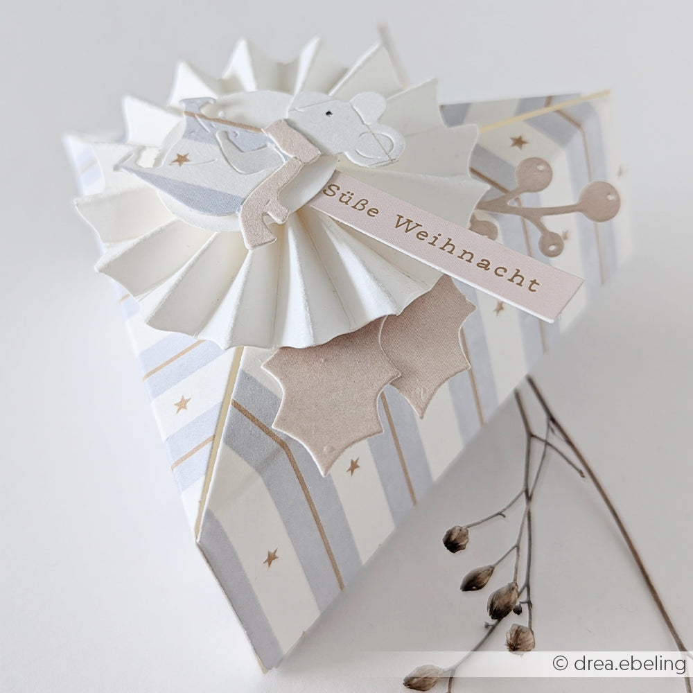 Dreieckige Geschenkbox mit einem Design aus Aquarellstreifen, einem Bären, einer Papierrosette und der Aufschrift Süße Weihnacht.