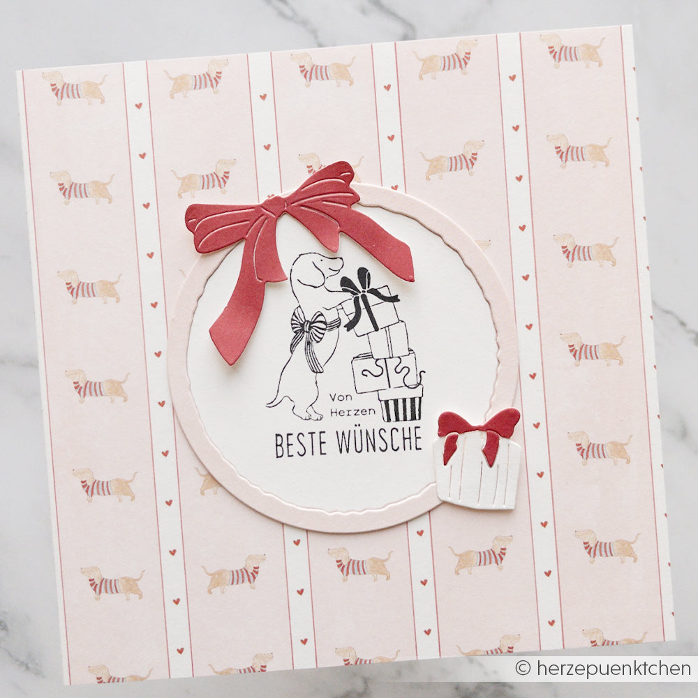 Grußkarte mit einem Streifenmuster im Dackel-Design, auf der ein Hund mit Geschenken abgebildet ist und die einen deutschen Text enthält.