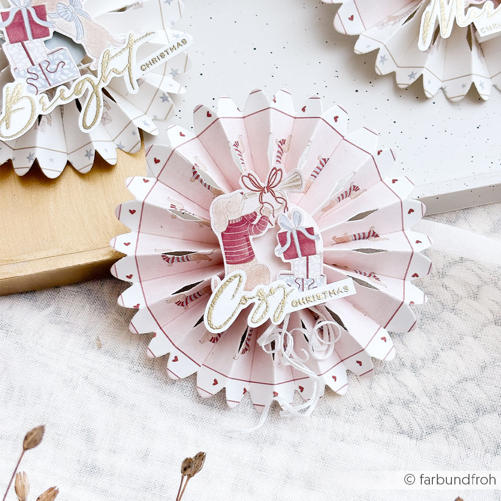 Weihnachtsbaumschmuck aus Papier in Form einer Rosette mit einem Hasen, dem Wort Joy und gemustertem Papier.
