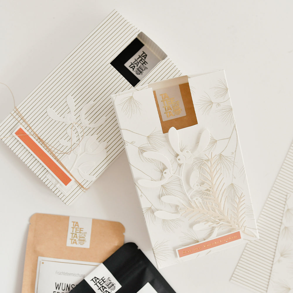 Zwei weiße Schachteln und zwei kleine Tütchen mit TaTee-Teeprodukten, die neben Designpapier ausgestellt sind.