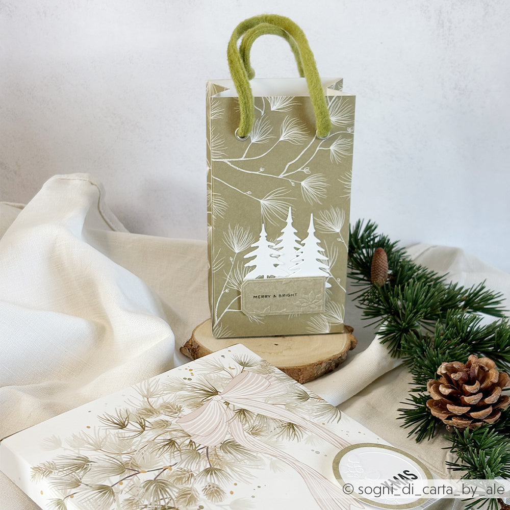 Olivgrüne Geschenktüte mit Papier mit weißem Kiefernmotiv und einem Merry & Bright-Anhänger.