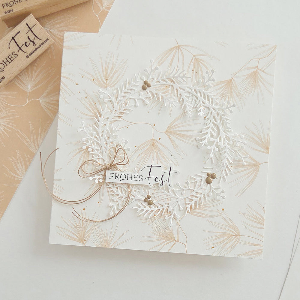 Quadratische Karte aus Designpapier mit einem weißen, ausgestanzten Kranz und einem Frohes Fest-Anhänger, der mit einer Schnur befestigt ist.