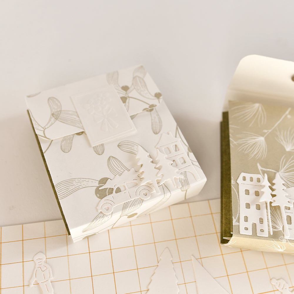 Zwei quadratische Geschenkboxen, umhüllt mit Designpapier „Misteln Sanftgrün“, verziert mit weißen und goldenen Motiven von Weihnachtsbäumen, Autos und weihnachtlichem Grün.