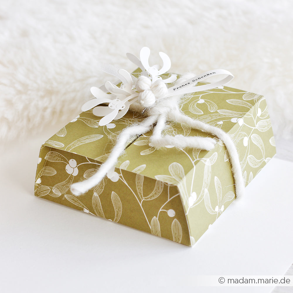Olivgrüne Geschenkbox mit einem Mistelzweig auf zartgrünem Designpapier, mit einer Schnur und einem Anhänger verschnürt.