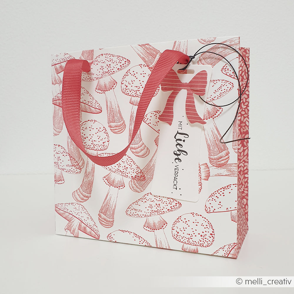 Geschenktüte mit rotem Pilzmuster, rotem Band als Tragegriff und einem Anhänger mit der Aufschrift Mit Liebe verpackt.