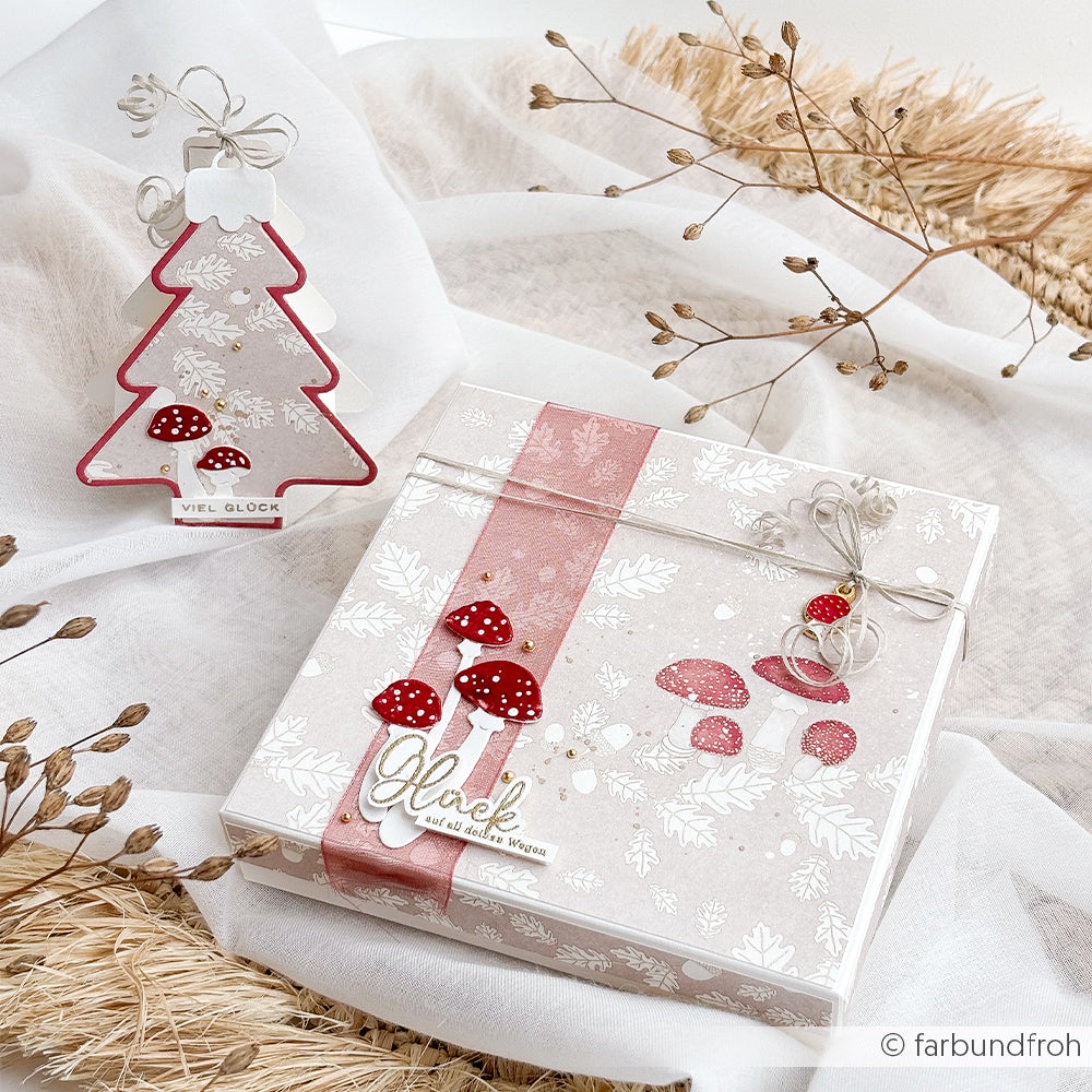 Eine quadratische Geschenkbox und ein Anhänger in Form eines Weihnachtsbaums mit dekorativen Pilzmotiven und dem deutschen Wort „Glück“.