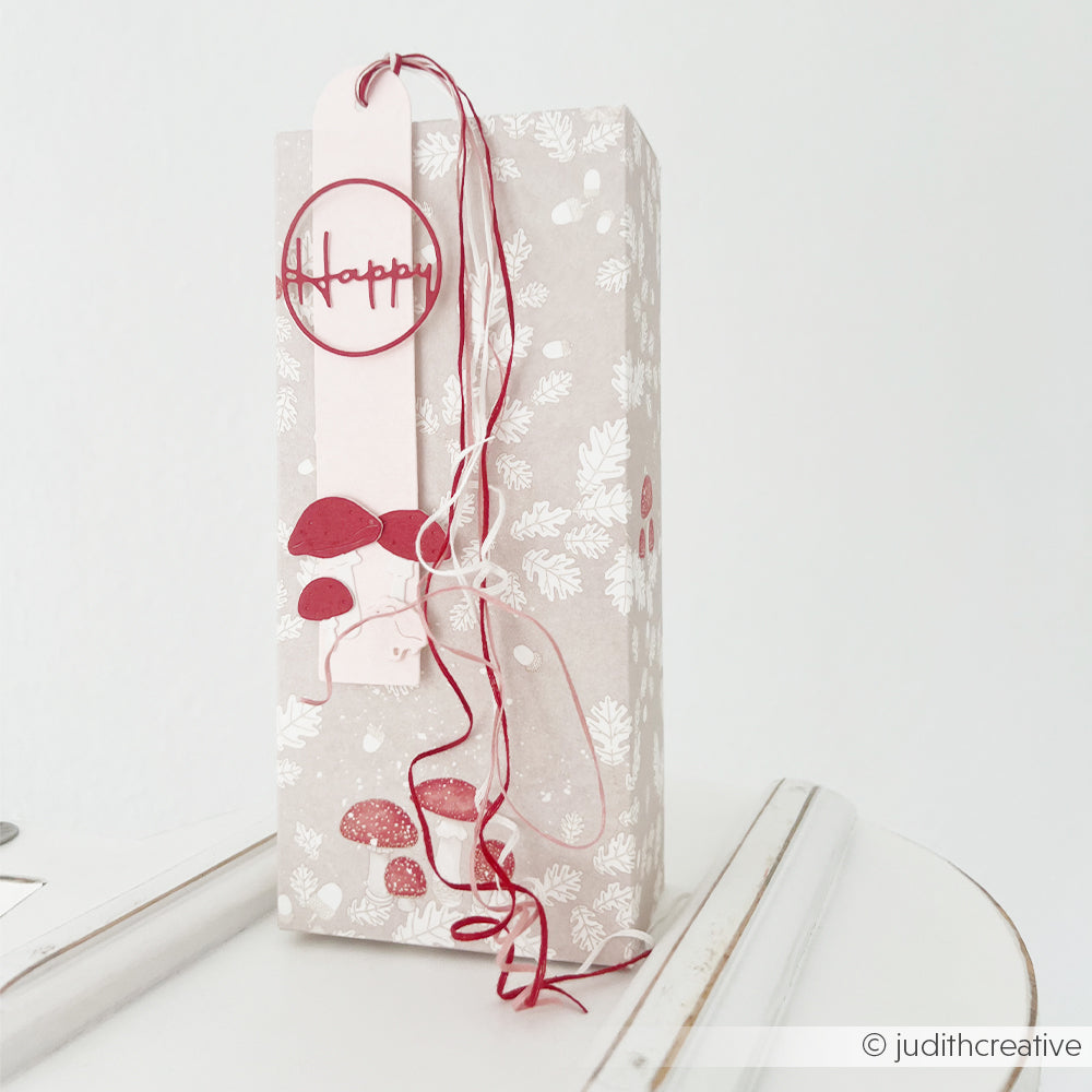 Hohe Geschenkbox mit Pilzmotiven, einem Happy-Anhänger und dekorativem Designpapier.