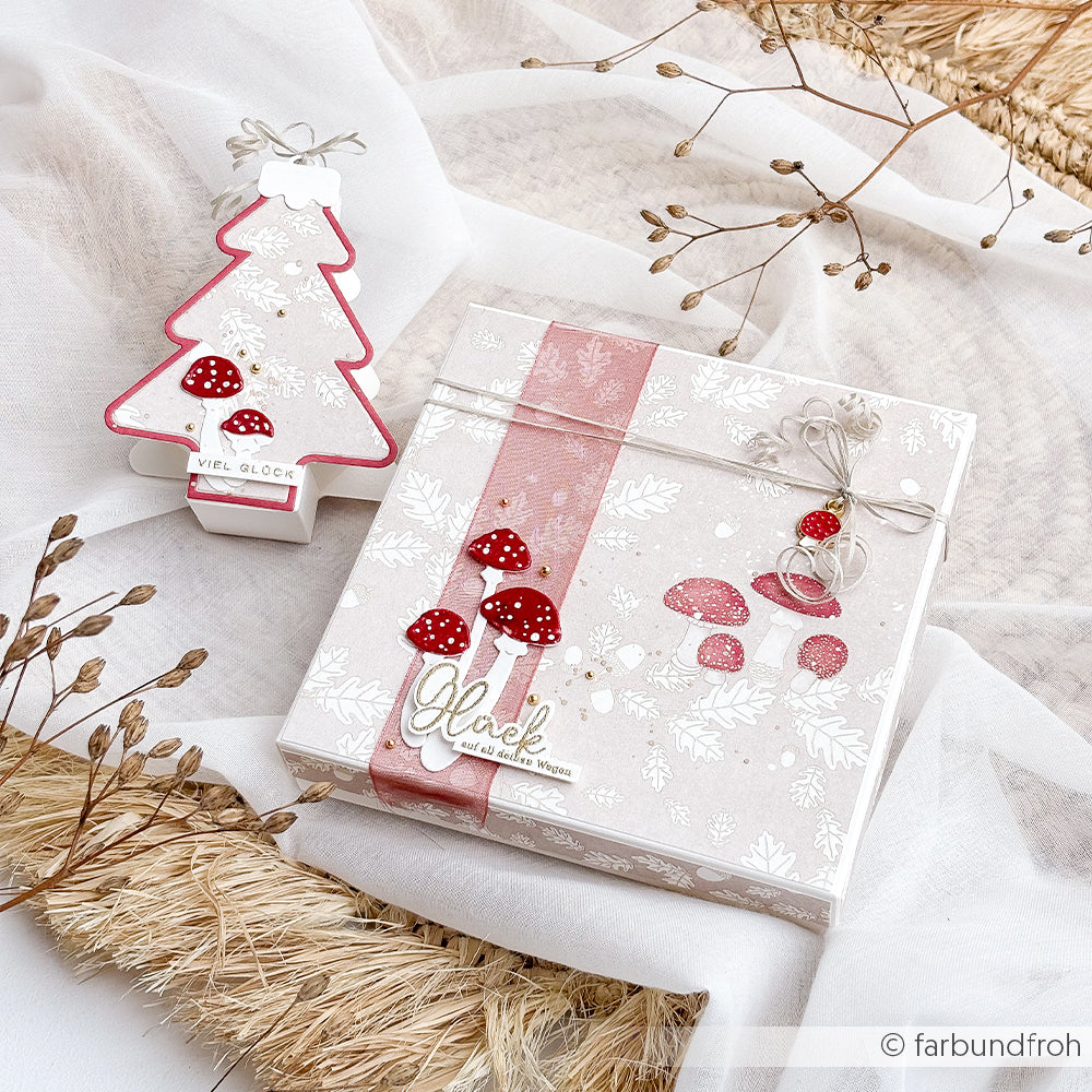 Eine Geschenkbox und ein baumförmiger Christbaumschmuck, verpackt in Designpapier mit roten Pilzmotiven und dem deutschen Wort „Glück“.