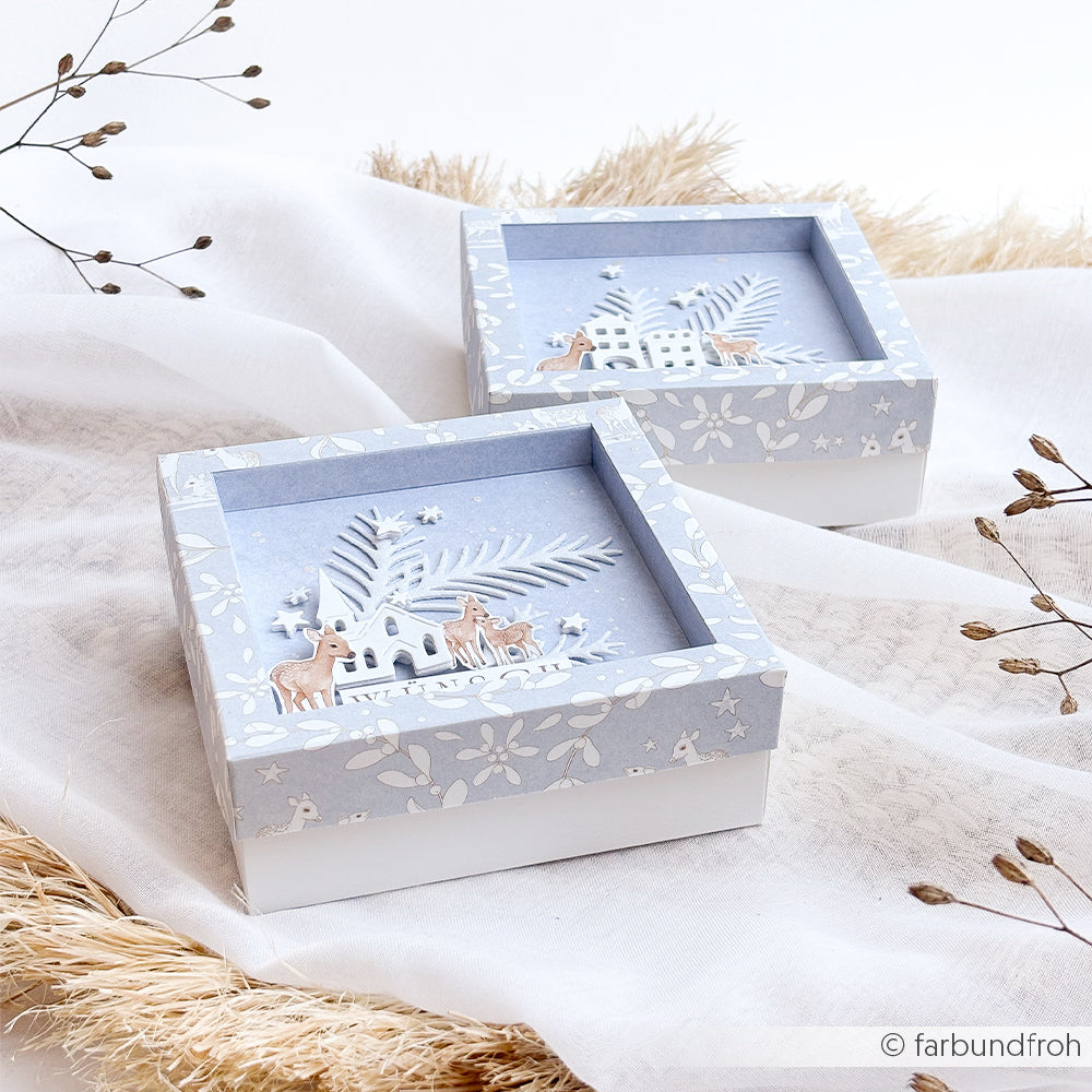 Zwei quadratische Geschenkboxen mit einer mehrschichtigen Winterlandschaft mit Rehen und einem Haus auf dekorativem Designpapier.