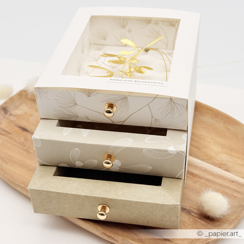 Eine Geschenkbox mit drei Schubladen in Eschengrün mit goldenem Mistelzweig und der Aufschrift Fröhliche Weihnachten.