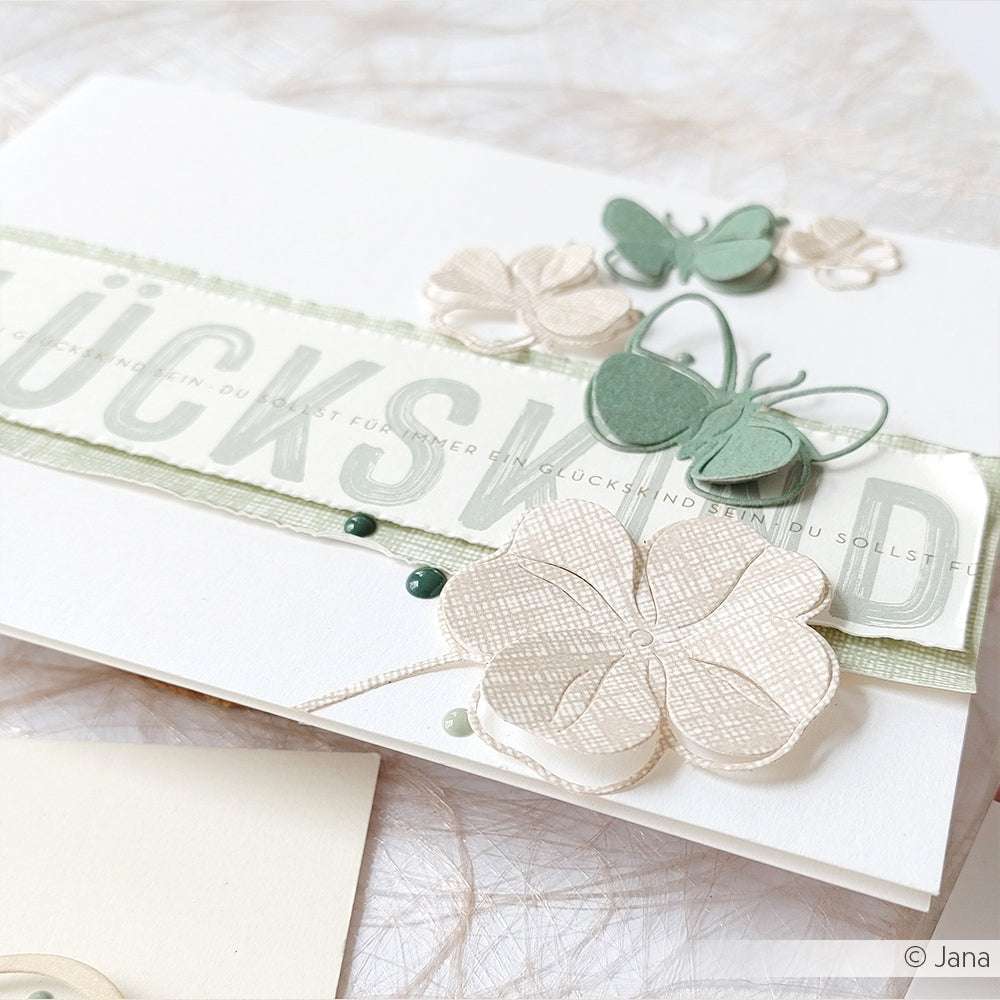 Handgefertigte Karte mit geschichteten Schmetterlingen und Blumen in zartem Grün und Beige, gefertigt aus Alexandra Renke Designpapier Leinen Schlamm hell-ideal für Buchbinderei oder Papierprojekte. Ein Teil des Textes verleiht der Karte eine zarte Note.