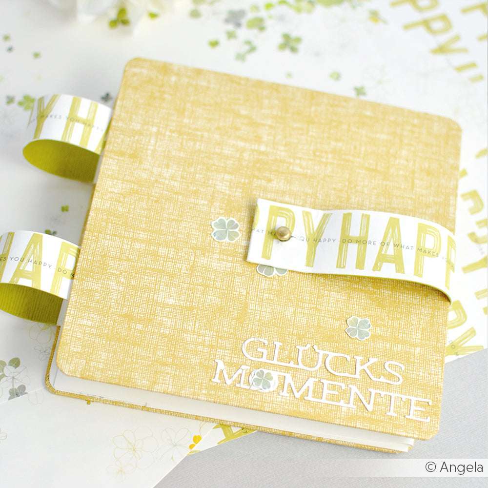 Ein gelbes Scrapbook von Alexandra Renke mit dem Text "GLÜCKSMOMENTE" und einem weiß-grünen Designpapier Leinen Honiggelb-Streifen - ideal für kreative Papierprojekte und buchbinderische Inspirationen.