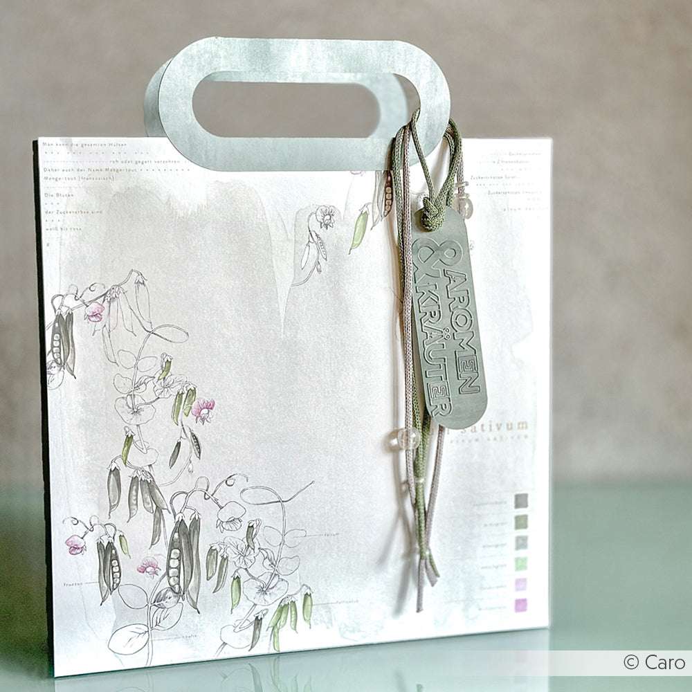 Weiße Geschenktüte von Alexandra Renke mit einem floralen Motiv auf hochwertigem Designpapier, verziert mit einem grünen Band und einem Anhänger mit der Aufschrift Aromen & Kräuter", ausgestellt auf einer hellen Fläche.