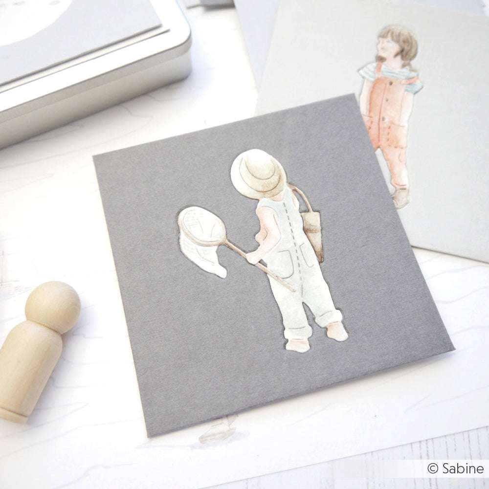 Alexandra Renke aquarellierte Illustration eines Kindes mit Hut und Netz auf grauem Karton, mit Designpapieren und einer Holzpuppe in der Nähe.