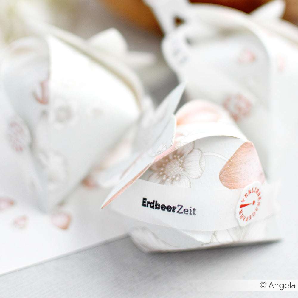 Nahaufnahme einer gefalteten, floral gemusterten Papierschachtel von Alexandra Renke, hergestellt aus Designpapieren und geformt mit einer zarten Motivstanze, beschriftet mit "ErdbeerZeit" und ausgestellt auf einer hellen Fläche.