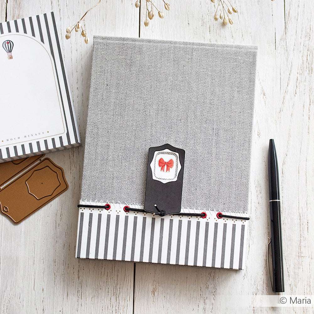 Ein graues ☆ Notizbuch mit gestreiften Buchleinen-Akzenten, einem Geschenkanhänger und einem schwarzen Stift auf einer weißen Holzoberfläche - ideal für diejenigen, die die hochwertige Buchbinden-Handwerkskunst von ☆ schätzen.