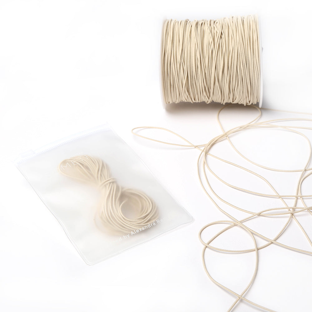 Eine Spule beigefarbenes Elastikband von Alexandra Renke mit einem kleinen Bündel in einem durchsichtigen Beutel, ideal für Bastelprojekte, dargestellt auf weißem Hintergrund.