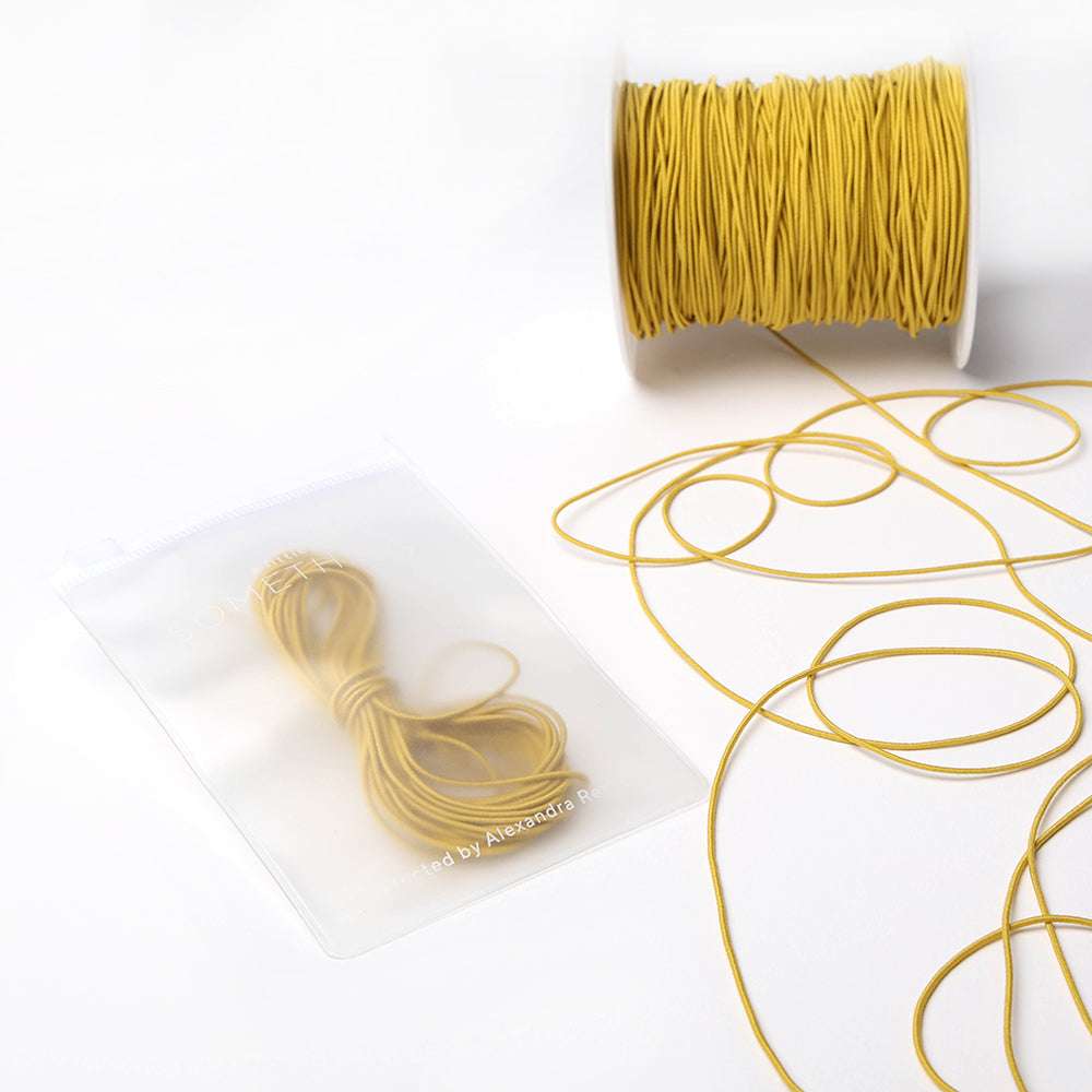 Eine Spule und ein Bündel dünnes gelbes Alexandra Renke Elastikband, mit einem Bündel in einem durchsichtigen Plastikbeutel auf weißem Hintergrund - perfekt für jedes Bastelprojekt.