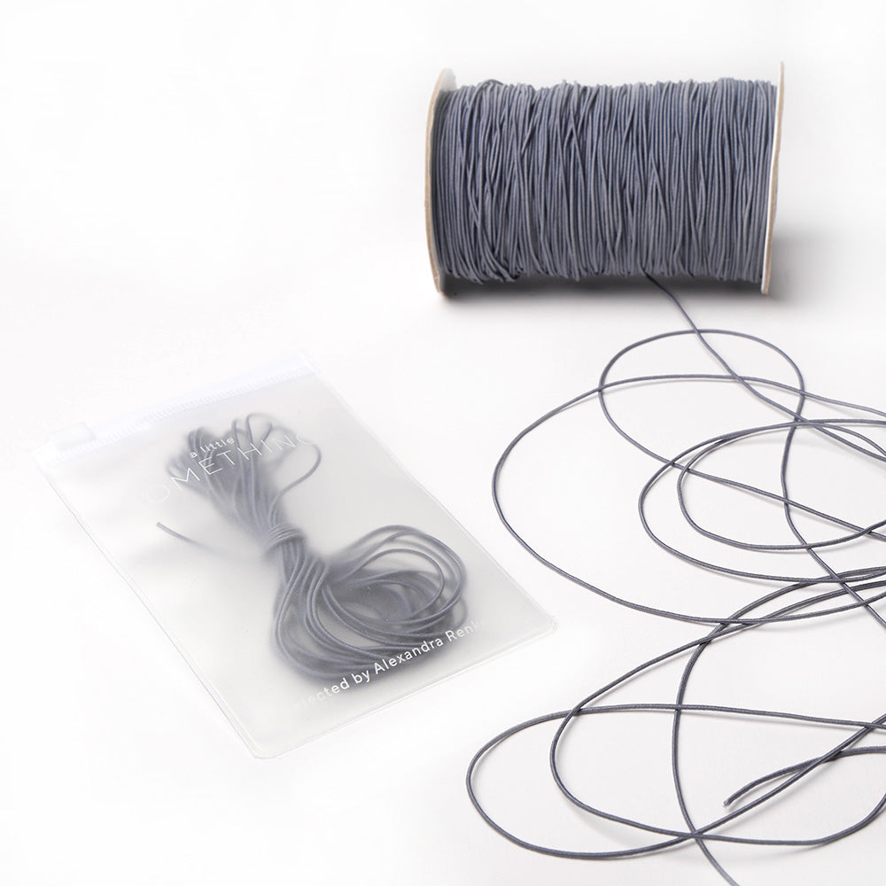 Graue Spule Alexandra Renke Elastikband mit loser Kordel daneben; aufgerollte Gummibänder in einem Klarsichtbeutel - ideal für Ihr nächstes Bastelprojekt.