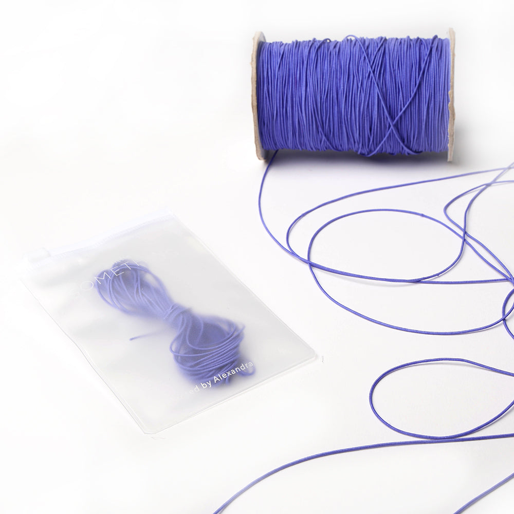 Eine Spule und ein Bündel lila Elastikband von Alexandra Renke, verpackt in einer durchsichtigen Plastiktüte auf weißem Hintergrund - perfekt für Ihr nächstes Bastelprojekt.