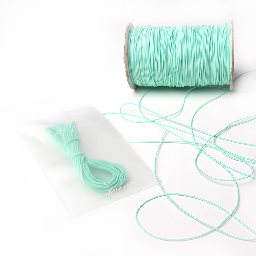 Eine Spule hellblaues Alexandra Renke Elastikband mit einem losen Bündel, ideal für jedes Bastelprojekt, kommt in einem wiederverschließbaren Beutel auf weißem Hintergrund.