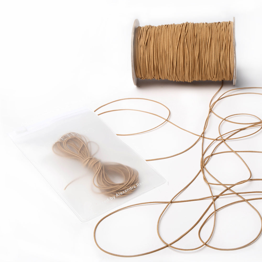 Eine Spule und ein Bündel hellbraunes Elastikband von Alexandra Renke, mit etwas loser Kordel auf einer weißen Fläche - ideal für Ihr nächstes Bastelprojekt.