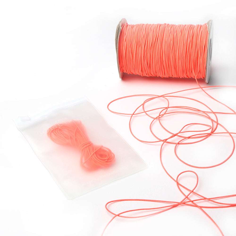 Eine Spule dünnes korallenfarbenes Elastikband von Alexandra Renke mit etwas loser Kordel und einem kleinen Bündel in einem durchsichtigen Plastikbeutel - ideal für Bastelprojekte.