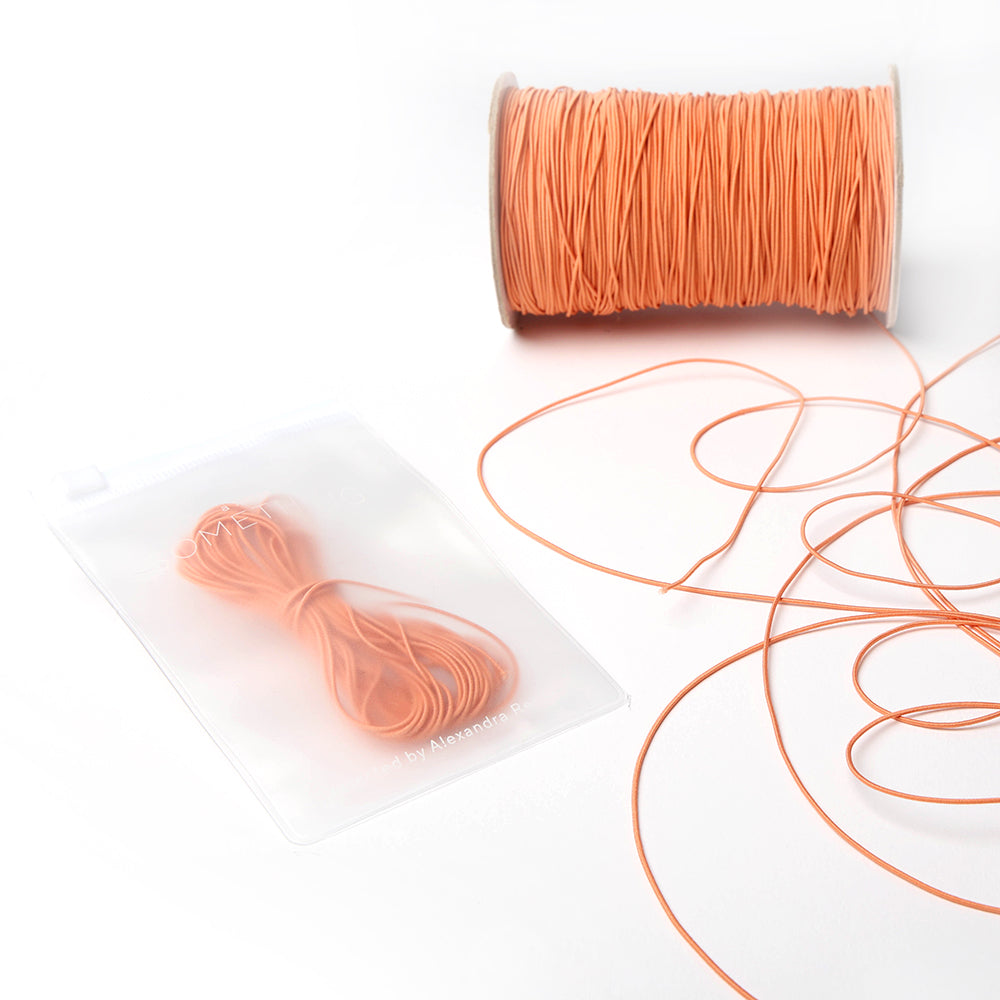 Das pfirsichfarbene Elastikband von Alexandra Renke ist fein säuberlich auf eine Spule gewickelt, während die Gummibänder in einem durchsichtigen Beutel - perfekt für jedes Bastelprojekt - auf einer weißen Fläche präsentiert werden.