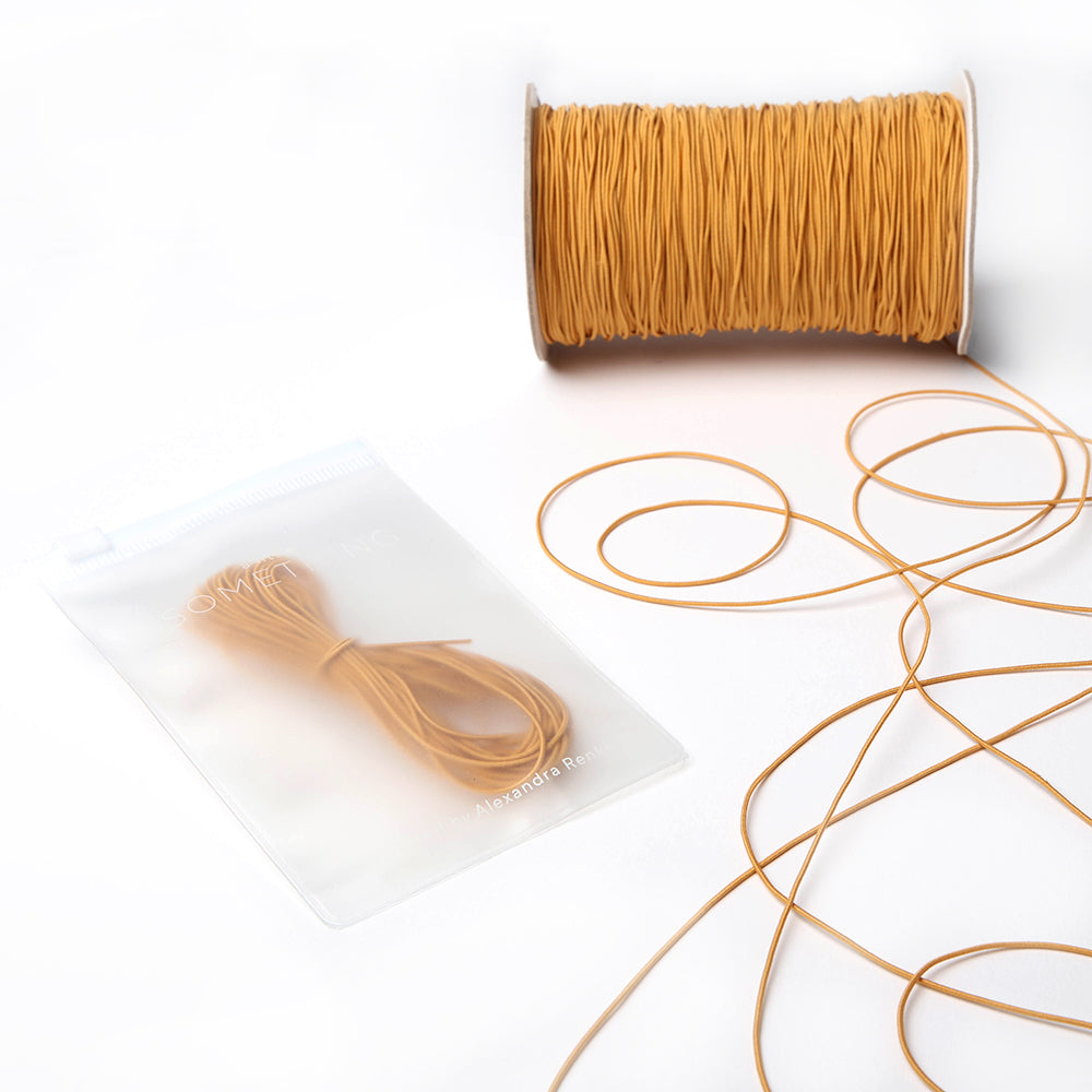 Eine Spule und ein kleines Bündel hellbraunes Elastikband von Alexandra Renke, mit Kordel locker auf einer weißen Fläche angeordnet - genau richtig für Ihr nächstes Bastelprojekt.