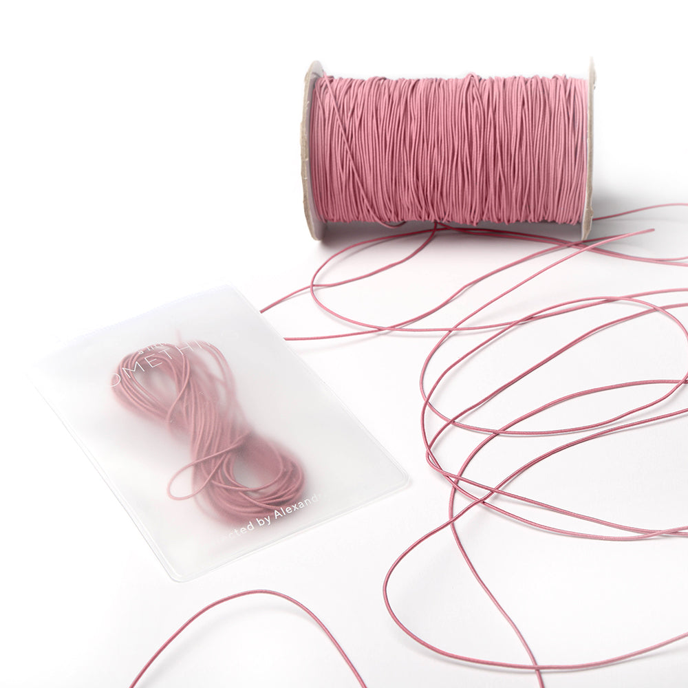 Eine Spule und ein Bündel rosafarbenes Elastikband von Alexandra Renke, mit etwas loser Kordel auf einer weißen Fläche - perfekt für Ihr nächstes Bastelprojekt.