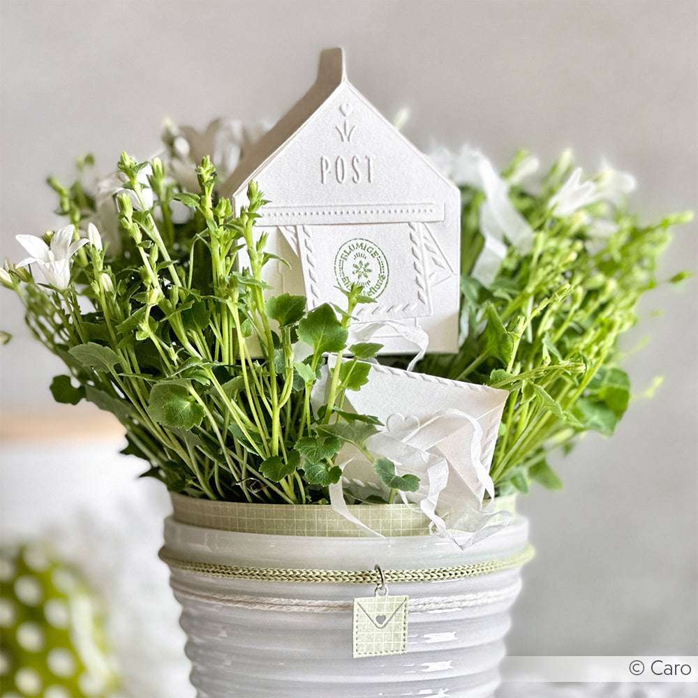 Weiße Blumen in einem weißen Topf mit einem dekorativen Papierhaus und einem grün-weiß-silbernen Anhänger, der mit der Schneide- und Prägefoldertechnik von Alexandra Renke erstellt wurde.