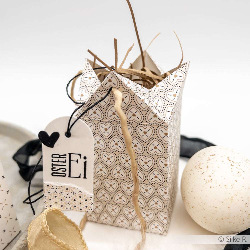 Dekorative Geschenkschachtel von Alexandra Renke mit Herzmotiven, Strohfüllung und einem "Oster Ei"-Anhänger, hergestellt mit Motivstanze Schachtel, ausgestellt neben einem weiß gesprenkelten Ei.