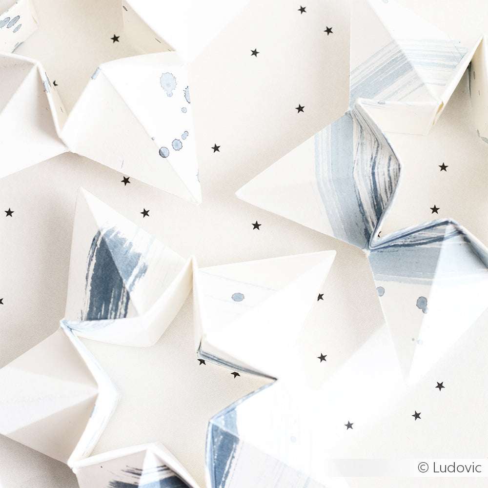 Weiße und blaue Origami-Sterne aus Designpapier von Alexandra Renke ruhen auf einem weißen Hintergrund mit schwarzen Stern-Akzenten - ideal, um die Kreationen der Motivstanze Doppel-Faltstern zu präsentieren.